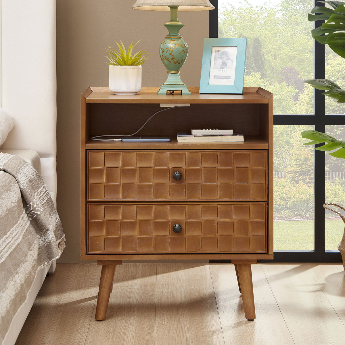 Light brown nightstand beside a bed, with two drawers, open shelf, and charging cable