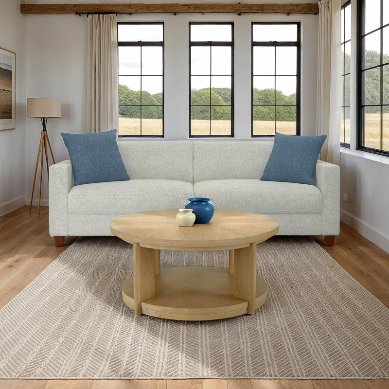 Round light-brown wood coffee table with lower shelf in front of a sofa