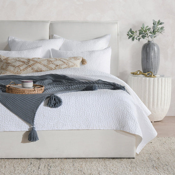 Neutral quilt on bed with multiple pillows and a dark gray accent throw.