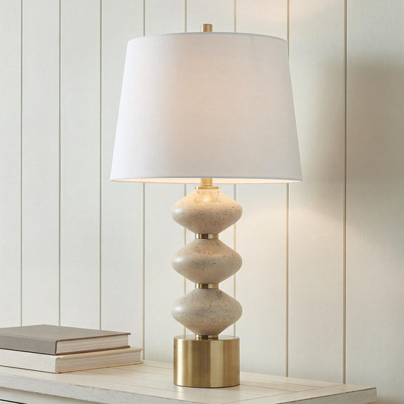 Beige table lamp with white fabric shade and brass base over stacked speckled bodies