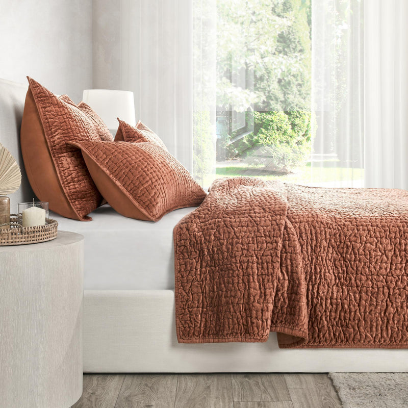 Rust velvet quilt set with textured quilt and matching decorative pillows on sofa.