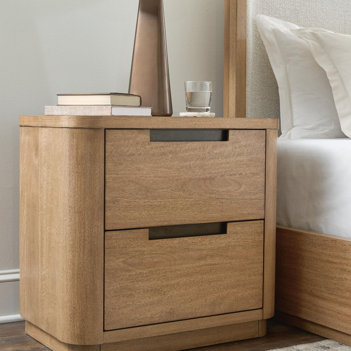 Light brown two-drawer night stand beside bed, featuring black bar pull handles.