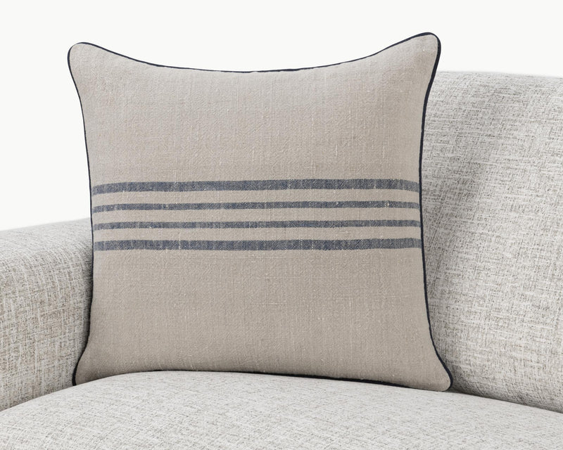 Natural linen pillow with blue stripe band and dark piping, shown on a sofa.
