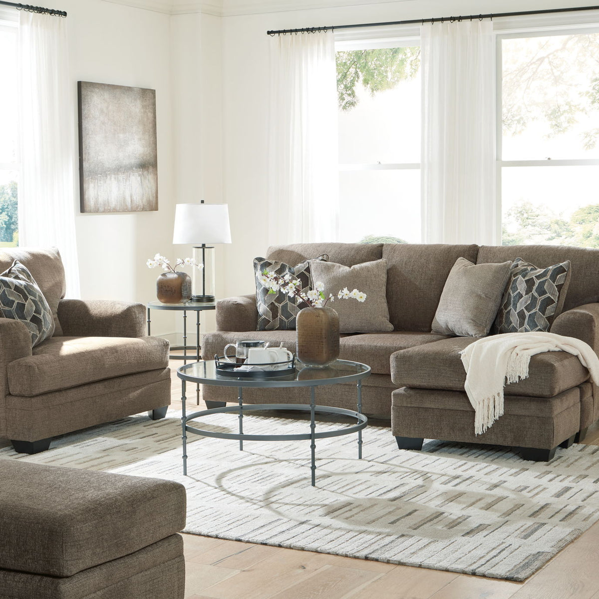 Taupe fabric sofa set with chair, ottoman, and patterned throw pillows in a bright living room.