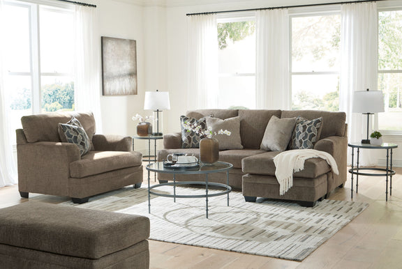 Taupe fabric sofa set with chair, ottoman, and patterned throw pillows in a bright living room.