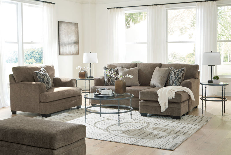 Taupe fabric sofa set with chair, ottoman, and patterned throw pillows in a bright living room.