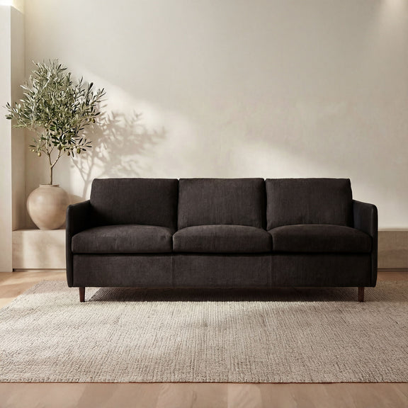 Upholstered sofa in dark warm black with three seat cushions, boxy back, tapered legs.