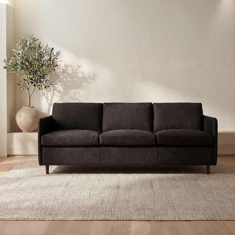 Upholstered sofa in dark warm black with three seat cushions, boxy back, tapered legs.