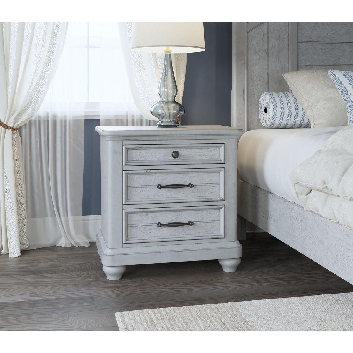 Gray wood 3-drawer nightstand with metal pulls and turned legs beside bed.