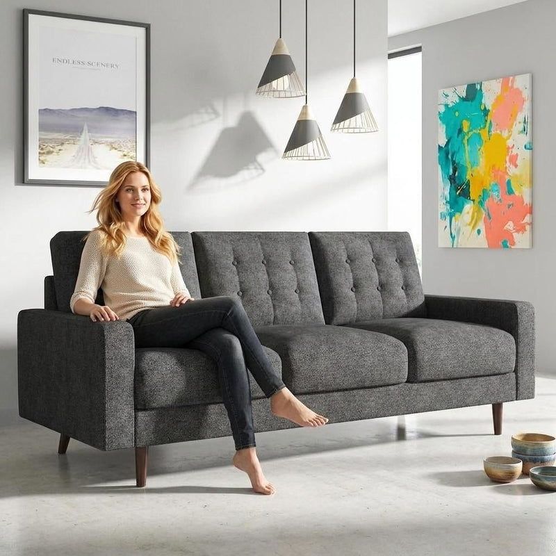 Dark gray three-seat tufted sofa with wide arms and wood legs, shown with one seated person