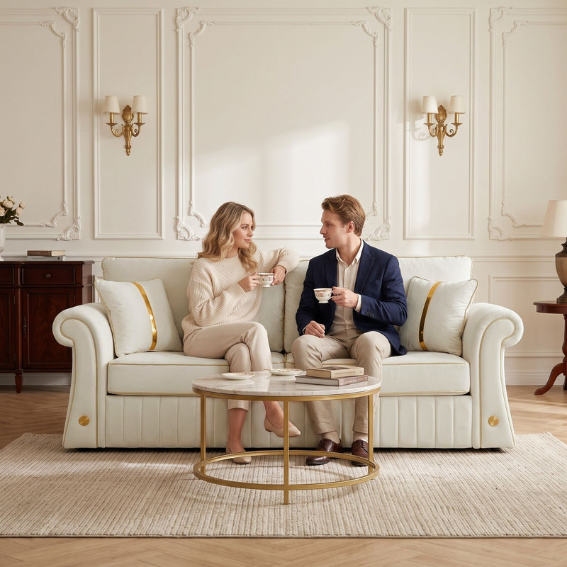 Cream three-seat sofa with rolled arms and gold-trimmed pillows, shown with two seated people