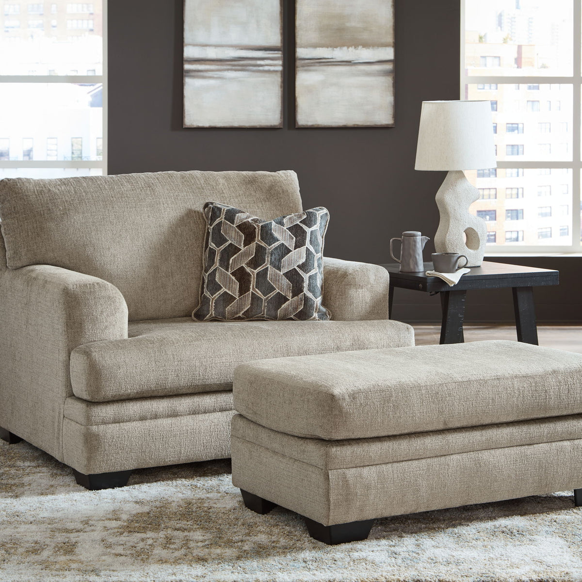 Taupe fabric loveseat with square arms and matching ottoman, styled with patterned throw pillow.