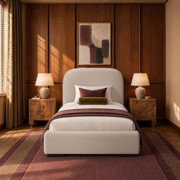 Beige upholstered bed with rounded headboard, pillows, and blanket in a bedroom setting