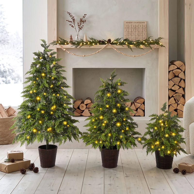 Three green artificial Christmas trees with cascading branches, warm LED lights, and woven pots.
