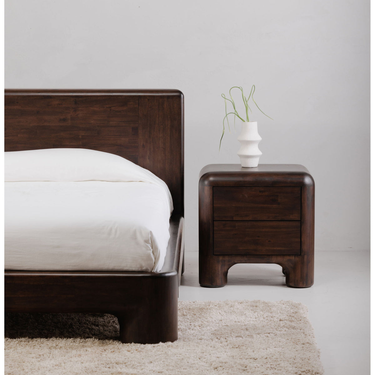 Dark brown wood nightstand with single drawer and rounded legs, next to bed