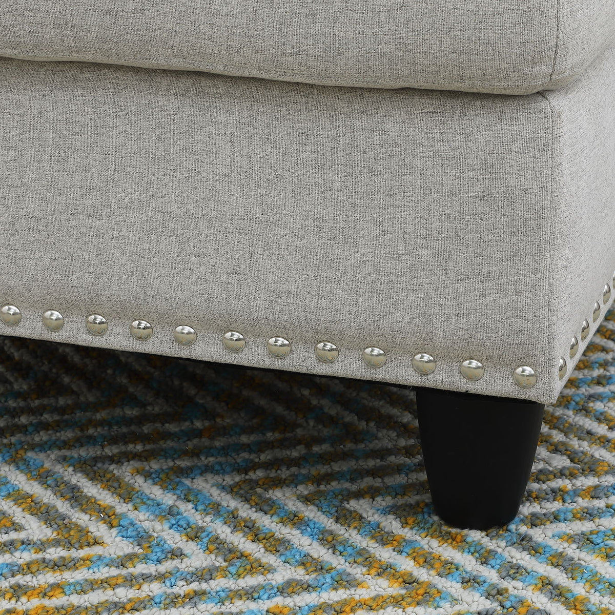Gray sofa corner with nailhead trim and dark legs, shown over patterned rug