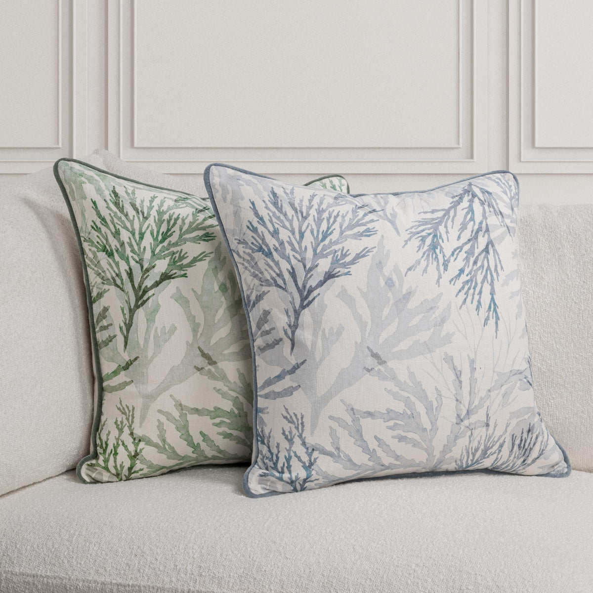 Two botanical decorative throw pillows on a sofa: green and blue designs with matching piping