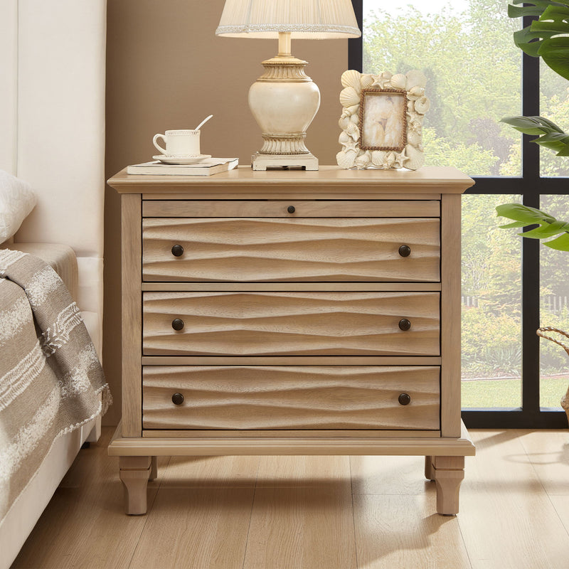 Light brown nightstand with three sculpted drawers, shown beside a bed